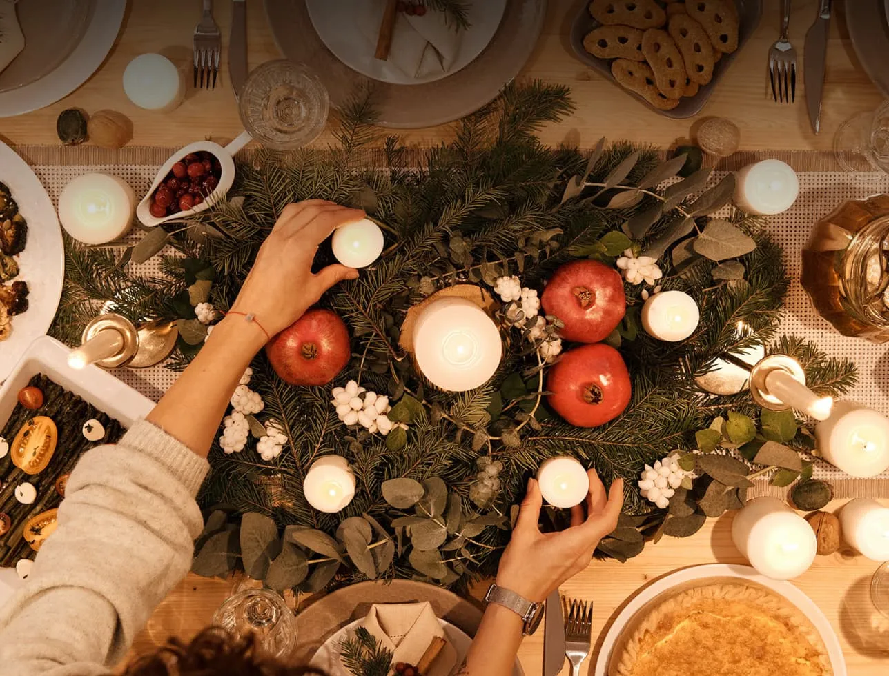 Top-down view of a festive table centerpiece with evergreen branches, pomegranates, white berries, and lit candles being arranged by a person.